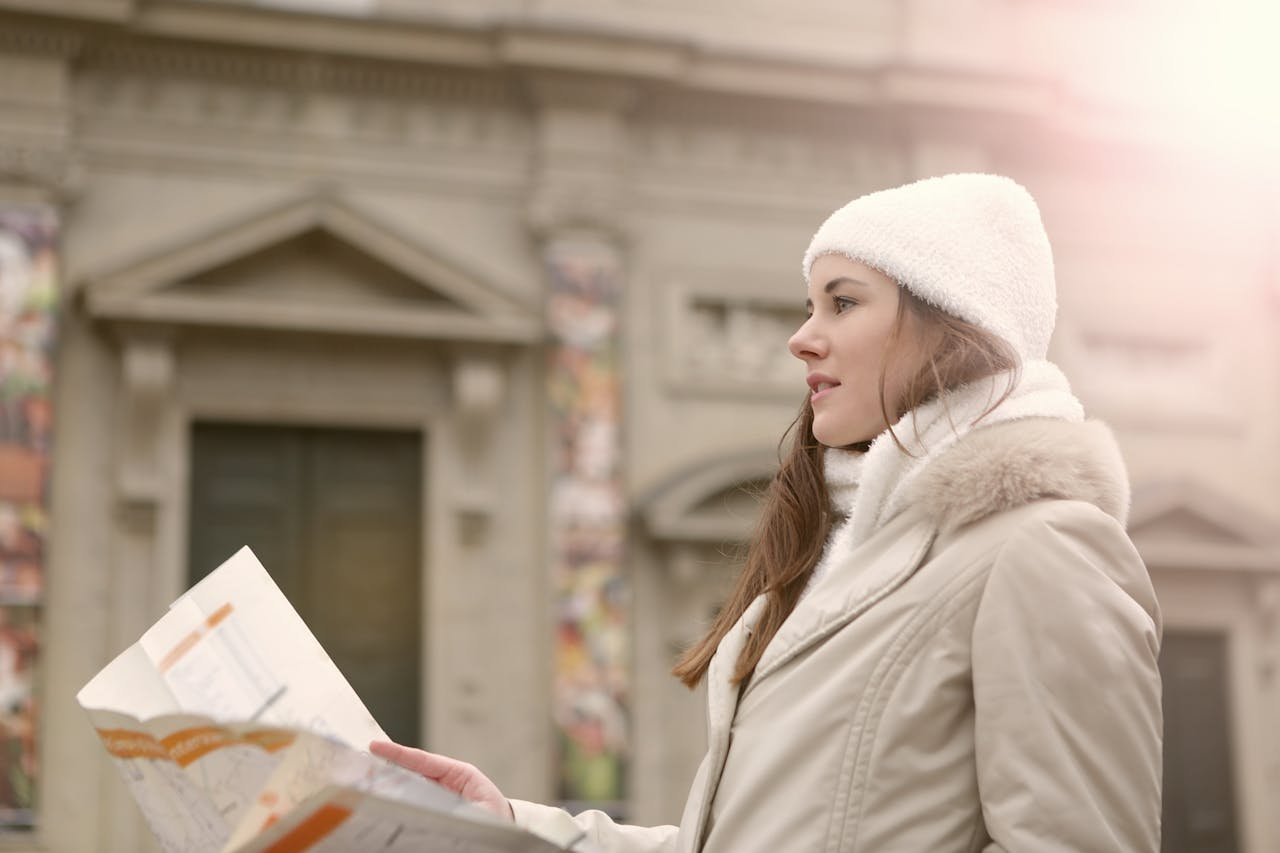 Side view of female traveler in warm clothes and head wear studying map while standing near antique building