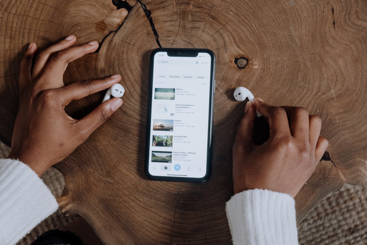 Hands interacting with smartphone and earbuds on a rustic wooden table, highlighting modern technology.