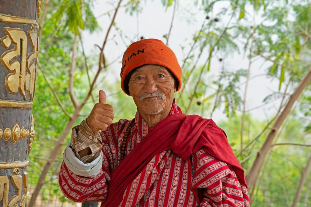 A senior Bhutanese man wearing traditional red clothing and an orange hat gives a thumbs up outdoors.